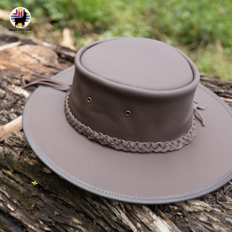 May include: A taupe-colored leather hat with a wide brim and a braided band. The hat features a rounded crown and decorative fringe. The brim has a gray trim. The hat is resting on a weathered log.