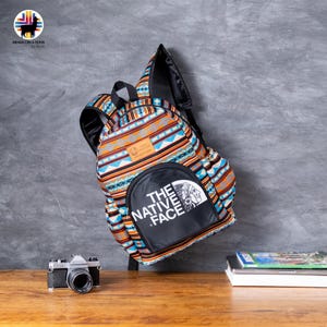 May include: A colorful backpack with a geometric pattern in orange, blue, and white. The backpack has a black front pocket with the text "THE NATIVE FACE" and a Native American graphic. A vintage camera and books are on the table.