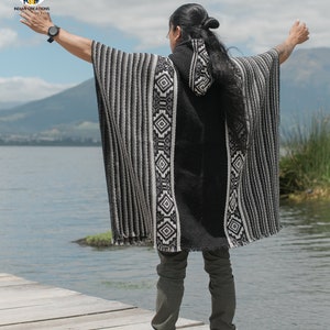 Pode incluir: Uma pessoa vestindo um poncho com capuz listrado de preto e branco com padr&otilde;es geom&eacute;tricos. O poncho &eacute; feito de l&atilde; de alpaca e tem um design tradicional. A pessoa est&aacute; em p&eacute; em um p&iacute;er de madeira com vista para um lago.