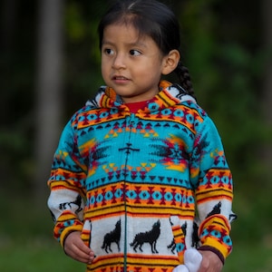 May include: A young person wearing a colorful hooded sweater with a geometric pattern and black wolf silhouettes. The sweater has a zipper closure and is made of a soft, knitted fabric. The person is holding a white stuffed animal.