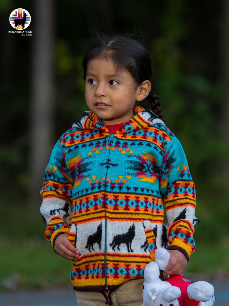 May include: A young person wearing a colorful hooded sweater with a geometric pattern and black wolf silhouettes. The sweater has a zipper closure and is made of a soft, knitted fabric. The person is holding a white stuffed animal.