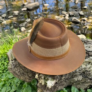 May include: A brown leather hat with a wide brim and a brown and tan band. A single feather is attached to the hat.