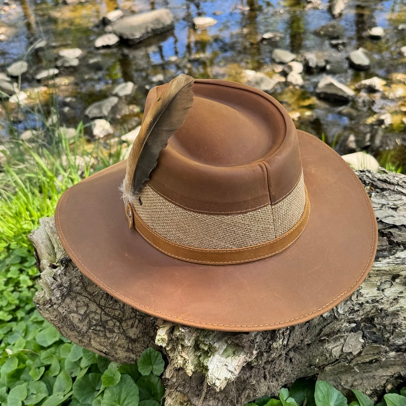 May include: A brown leather hat with a wide brim and a brown and tan band. A single feather is attached to the hat.