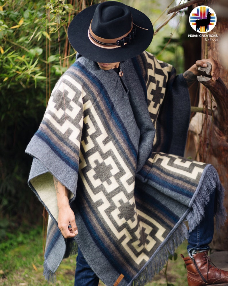 May include: A gray and white patterned poncho with fringe, worn by a person wearing a black fedora and blue jeans. The poncho has a geometric design with a brown and blue border.