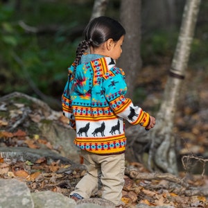 May include: A child wearing a turquoise, yellow and orange patterned hooded sweater with black wolf silhouettes. The sweater has a geometric design and is worn over khaki trousers.
