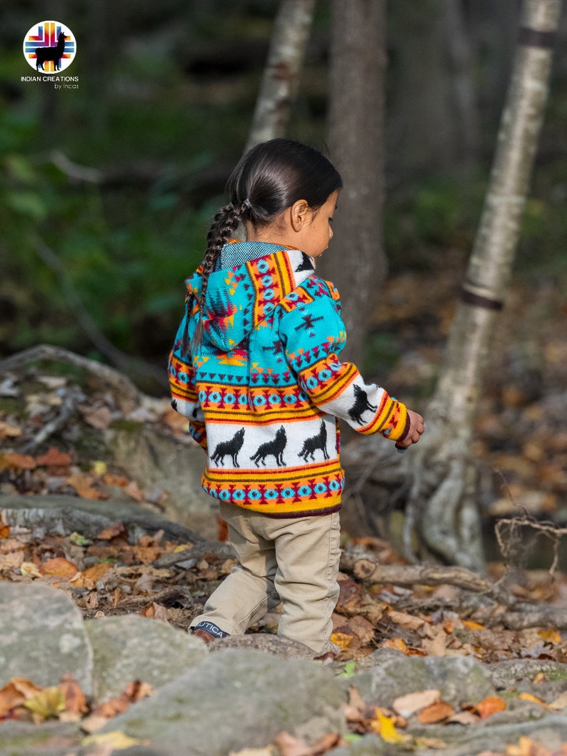 May include: A child wearing a turquoise, yellow, and orange patterned hooded sweater with black wolf silhouettes. The sweater has a geometric design and is worn over khaki pants.