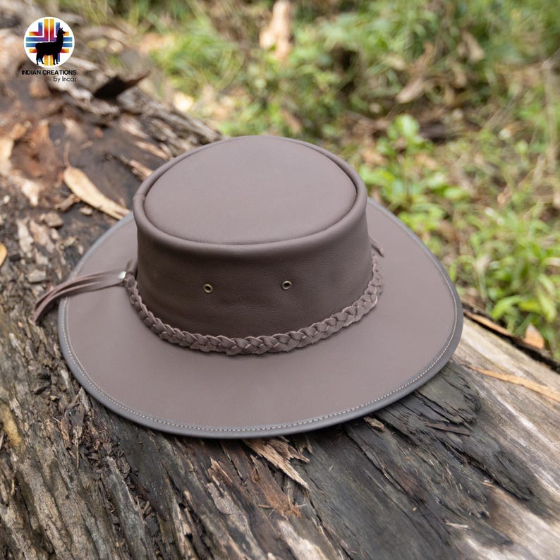 May include: A brown leather hat with a wide brim and a braided band around the crown. The hat has a rounded top and a small leather strap with a tassel. The hat is sitting on a weathered log.