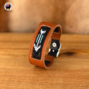 Pode incluir: Um bracelete de couro castanho com uma incrustação de couro preto que apresenta um design de contas brancas, pretas e turquesa. O bracelete tem um fecho de fivela.