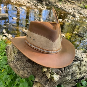 May include: A brown leather cowboy hat with a tan fabric band and a brown leather brim. The hat has a feather on the side.