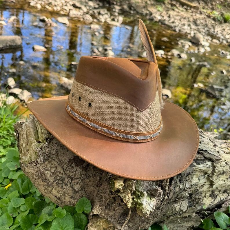 May include: A brown leather cowboy hat with a tan fabric band and a brown leather brim. The hat has a feather on the side.