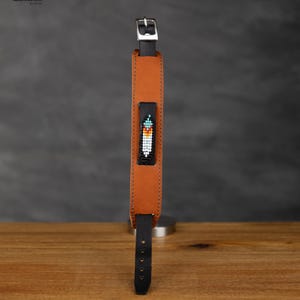 Può includere: Un cinturino da orologio in pelle marrone con fibbia nera e un design a piuma con perline. Il cinturino è su una superficie di legno.
