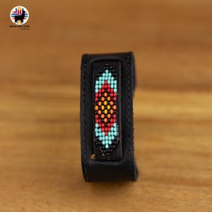 Può includere: Un bracciale a polsino in pelle nera con un motivo centrale di perline. Il motivo a perline presenta un design simmetrico con perline rosse, arancioni, gialle e turchesi. Il bracciale è mostrato su una superficie di legno.
