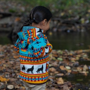 May include: A young person wearing a turquoise, orange, and white hooded sweater with a wolf pattern. The sweater has a colorful geometric design. The person is standing in a forest setting with fallen leaves on the ground.