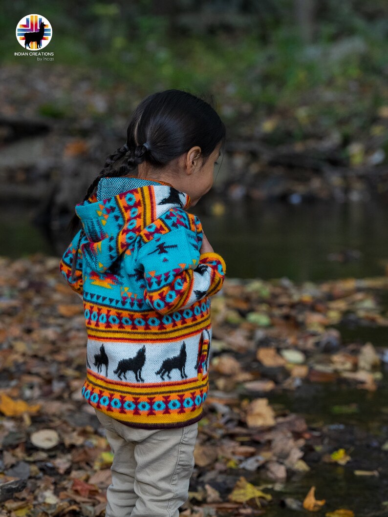 May include: A young person wearing a turquoise, orange, and white hooded sweater with a wolf pattern. The sweater has a colorful geometric design. The person is standing in a forest setting with fallen leaves on the ground.