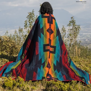 May include: A person wearing a colourful woven blanket with geometric patterns in shades of red, orange, blue, and green. The blanket is draped over the person's shoulders and is flowing in the wind. The person is standing on a grassy hill with a mountain range in the background. The text "INDIAN CREATIONS by Inca" is visible in the top right corner of the image.