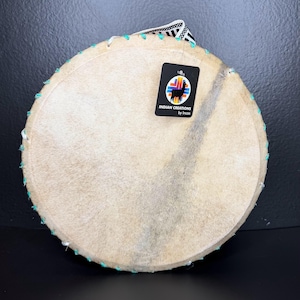 May include: A round, light-colored drum with a natural hide surface. The drum's edge is stitched with turquoise thread. A black tag with a colorful logo and the text "INDIAN CREATIONS by Incas" is attached to the drum.