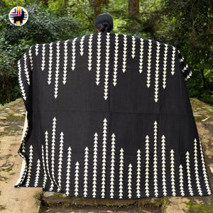 Puede incluir: Un poncho negro con un patrón repetitivo de triángulos blancos que apuntan hacia abajo. El poncho está drapeado sobre una persona, con un fondo verde natural. El poncho está hecho de un material suave y tejido. Las medidas del poncho no son visibles.