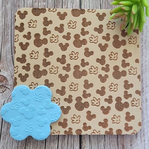 K&ouml;nnte beinhalten: Ein hellbraunes Quadrat mit einem sich wiederholenden Muster aus Mickey Mouse-K&ouml;pfen und -Handschuhen. Ein hellblaues, bl&uuml;tenf&ouml;rmiges Objekt mit demselben Mickey Mouse-Muster befindet sich in der linken unteren Ecke. Eine gr&uuml;ne Sukkulente befindet sich in der oberen rechten Ecke.
