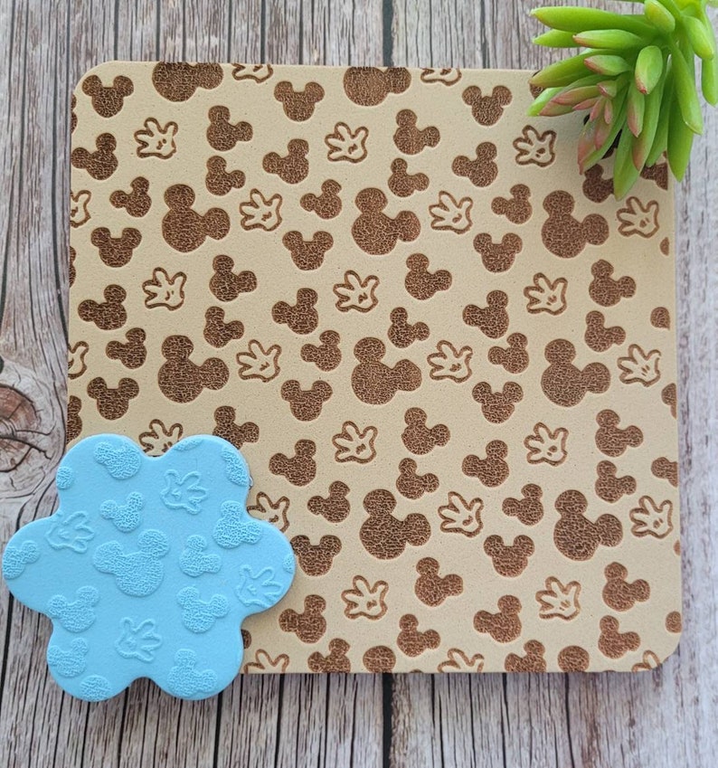 K&ouml;nnte beinhalten: Ein hellbraunes Quadrat mit einem sich wiederholenden Muster aus Mickey Mouse-K&ouml;pfen und -Handschuhen. Ein hellblaues, bl&uuml;tenf&ouml;rmiges Objekt mit demselben Mickey Mouse-Muster befindet sich in der linken unteren Ecke. Eine gr&uuml;ne Sukkulente befindet sich in der oberen rechten Ecke.