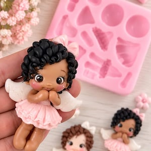 Puede incluir: Tres pequeñas figuras de ángeles de arcilla con cabello rizado oscuro o castaño, vistiendo vestidos con volantes rosas o blancos y alas blancas brillantes. Una figura se sostiene en la mano, mientras otras dos descansan junto a un molde de silicona rosa.