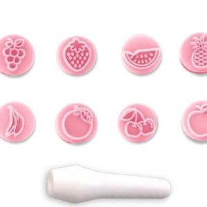 May include: A set of eight pink fruit-shaped stamps, including grapes, a strawberry, a watermelon slice, a pineapple, a banana, an orange, cherries, and an apple. A white handle is below the stamps. These stamps are likely for crafting or baking.
