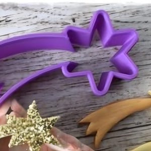 May include: A purple plastic star-shaped cookie cutter with a comet tail. The cutter is next to a baked star with a tail and other star-shaped cookies. The cookies are on a wooden surface.