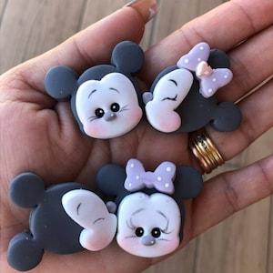 May include: Four grey Mickey Mouse and Minnie Mouse character-shaped embellishments. Each character has a white face, pink cheeks, and black eyes. Minnie Mouse characters have a light purple bow with white polka dots.
