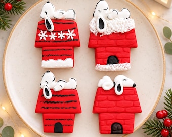 Puede incluir: Cuatro galletas decoradas con forma de casetas de perro rojas con figuras blancas de Snoopy encima. Cada galleta presenta detalles de glaseado distintos, incluyendo copos de nieve blancos, nieve texturizada y líneas negras.