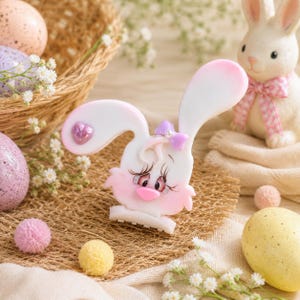 May include: A white and pink bunny-shaped hair clip featuring a small purple bow with a central pearl accent, a pink heart detail on one ear, and painted eyelashes. The clip has a smooth finish and a stylised, cartoon-like appearance.