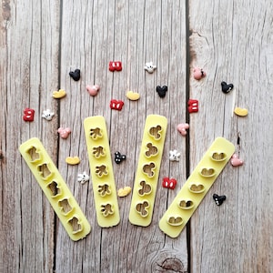 May include: Four yellow plastic cookie cutters with various shapes, including Mickey Mouse heads and gloves, are arranged on a wooden surface. Scattered around are small, colorful, Mickey Mouse-themed sprinkles in red, black, white, yellow, and pink.