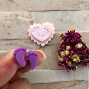 Puede incluir: Primer plano de dos pequeños amuletos morados con forma de pie. También se ve una decoración en forma de corazón rosa claro con huellas de bebé. Un pequeño ramo de flores secas con flores moradas y blancas está en el fondo.