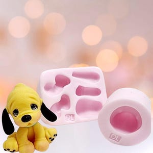 Może przedstawiać: Żółta figurka szczeniaka z czarnymi uszami i niebieską obrożą stoi obok różowych silikonowych foremek. Formy mają kształt części ciała szczeniaka. Tło jest miękko różowe ze światłami bokeh.
