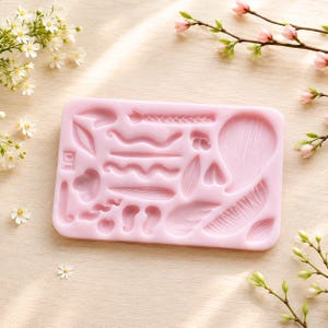 Puede incluir: Un molde de silicona rectangular de color rosa claro que presenta varias formas empotradas, incluyendo líneas onduladas, un patrón trenzado, formas de hojas y texturas de pétalos. El molde tiene una superficie lisa con múltiples cavidades.