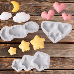 May include: A collection of silicone molds and plush shapes in the image. The molds are in the shape of clouds, stars, and hearts. The plush shapes include yellow stars, white clouds, pink hearts, and a yellow crescent moon. The items are arranged on a wooden surface.