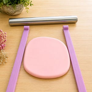 May include: A stainless steel rolling pin positioned horizontally above two parallel purple plastic guide sticks and a smooth, flat, circular piece of light pink dough centered between the guides on a light wood surface.