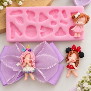 May include: A rectangular pink silicone mold with various small cavities, a lavender ribbon bow featuring a central unicorn-themed doll figure, and a separate small doll figure with brown hair and a red polka-dot bow headband.