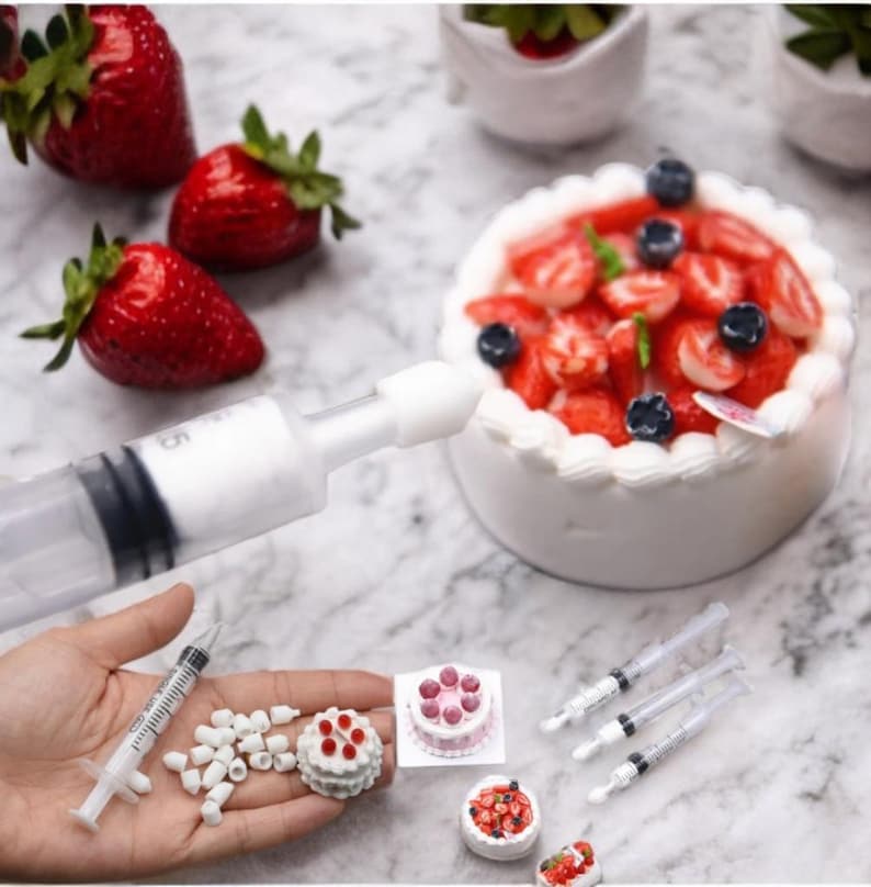 May include: A set of miniature cakes decorated with strawberries and blueberries, along with a syringe filled with white frosting and several empty syringes. The cakes are on a white surface with a larger cake in the background.