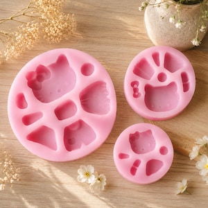 Puede incluir: Tres moldes redondos de silicona rosa que presentan formas hundidas de un personaje de gato de dibujos animados con un lazo, junto con varias cavidades pequeñas geométricas y de accesorios. Los moldes son lisos, flexibles y diseñados para moldear.
