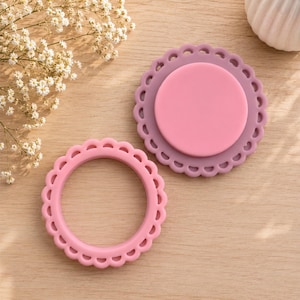 May include: Two circular silicone items with scalloped edges. One is a solid light pink ring, and the other is a mauve-colored circular base topped with a smooth, flat, light pink circular disc. Both items feature a matte finish and rounded edges.