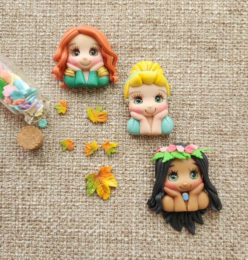 May include: Three handmade polymer clay figurines of Disney princesses. The princesses are Ariel, Cinderella, and Belle. Each figurine has a different hairstyle and facial expression. The figurines are all wearing different outfits.