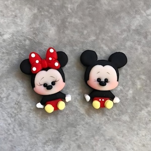 Peut inclure: Deux figurines de dessins animés, Mickey et Minnie Mouse, sont présentées. Minnie porte un nœud rouge à pois blancs. Les deux personnages ont des oreilles noires, des hauts noirs, des shorts rouges, des chaussures jaunes et des gants blancs. Les figurines sont sur une surface grise.