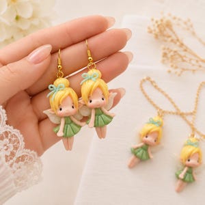 Puede incluir: Un par de pendientes colgantes con figuras de hadas en miniatura con cabello rubio recogido en mo&ntilde;os, lazos azul claro, vestidos verdes y alas blancas brillantes, suspendidos de ganchos de metal dorado, junto a dos dijes de hadas a juego.
