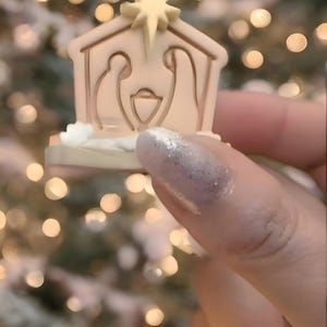 Puede incluir: Un pequeño adorno de belén beige con una estrella. Representa a María, José y el niño Jesús dentro de un establo. El fondo borroso presenta luces suaves, sugiriendo un árbol de Navidad.