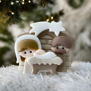 Puede incluir: Un belén navideño decorativo con María, José y el niño Jesús en un pesebre. Las figuras están en un establo con una estrella y un cometa. Las figuras son blancas, beige y marrones, sobre un fondo blanco.