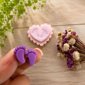Puede incluir: Un par de dijes morados con forma de huellas de bebé en primer plano, junto a un dije rosa claro en forma de corazón con huellas de bebé en relieve y un pequeño ramo de flores secas moradas y blancas al fondo.