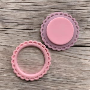May include: Two pink, scalloped-edge silicone molds. One is a complete circle with a raised center, and the other is a ring. The molds are in shades of pink and sit on a wooden surface. These are likely for crafting or baking.