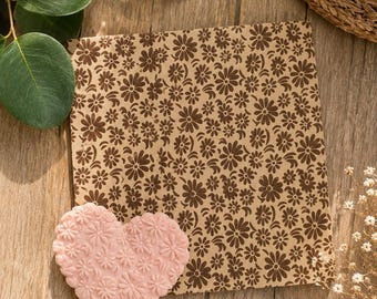 May include: A square piece of brown paper featuring a repeating dark brown floral pattern, accompanied by a heart-shaped object in light pink, which also displays a raised, repeating floral texture across its entire surface.