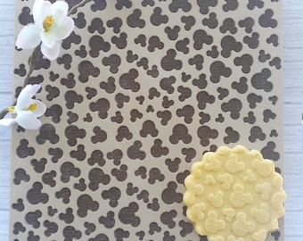 May include: A brown silicone baking mat with a repeating pattern of small Mickey Mouse heads. A yellow silicone cookie with the same pattern is on top of the mat.