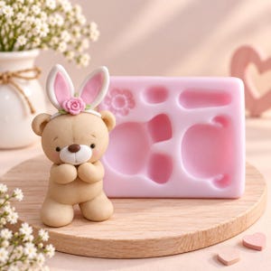 Puede incluir: Una figura de oso de color marrón claro que lleva una diadema blanca con orejas de conejo rosas y una pequeña rosa rosa, situada junto a un molde de silicona rosa rectangular con formas cóncavas para crear artículos decorativos.