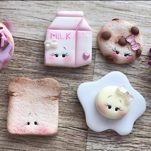 May include: A collection of miniature food-themed figurines, including a donut with purple frosting and sprinkles, a carton of milk, a cookie with chocolate chips, a slice of toast, and a fried egg. Each item has a cute face.